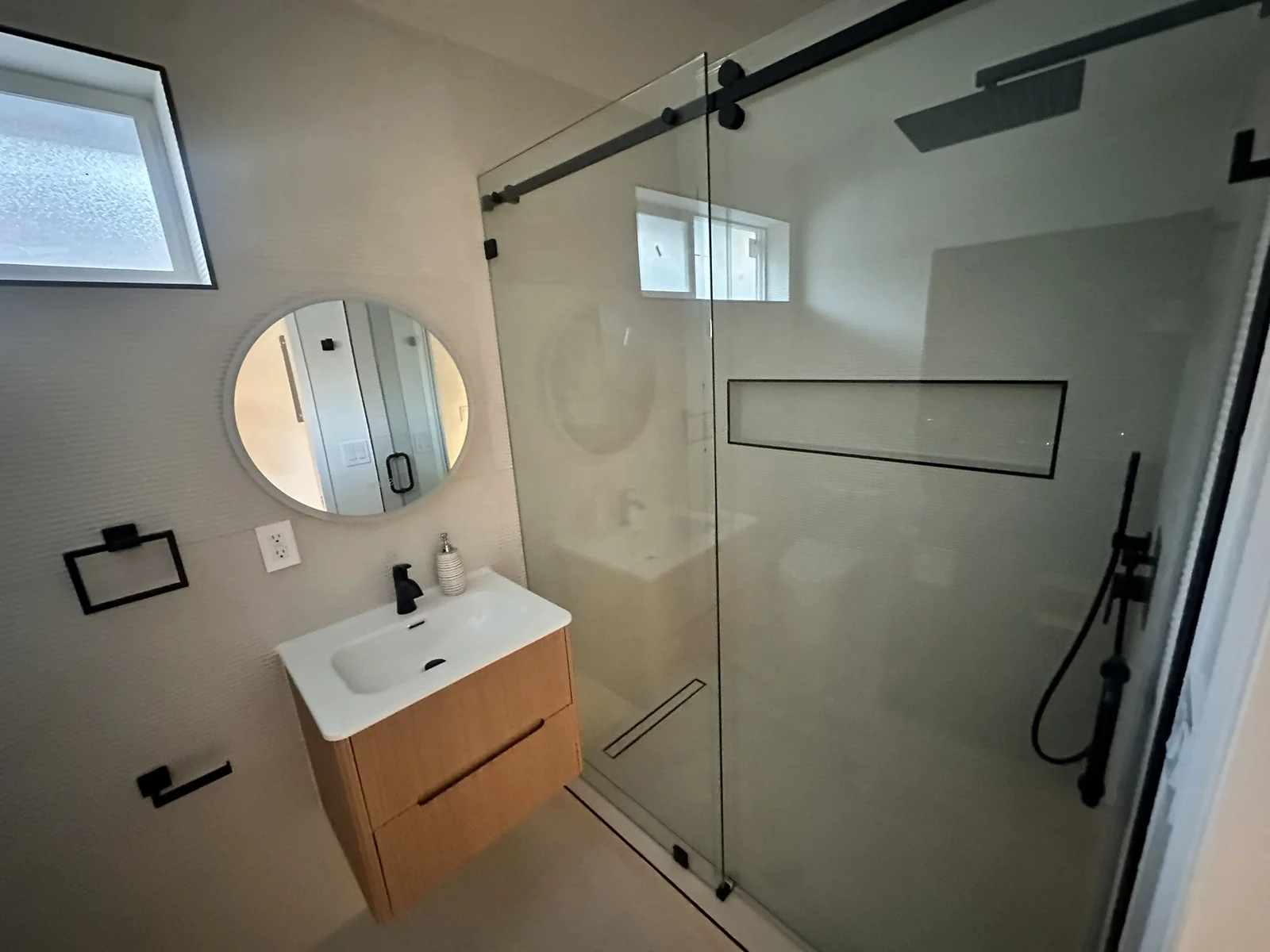 Compact bathroom with black-framed sliding glass shower, round mirror and wood vanity