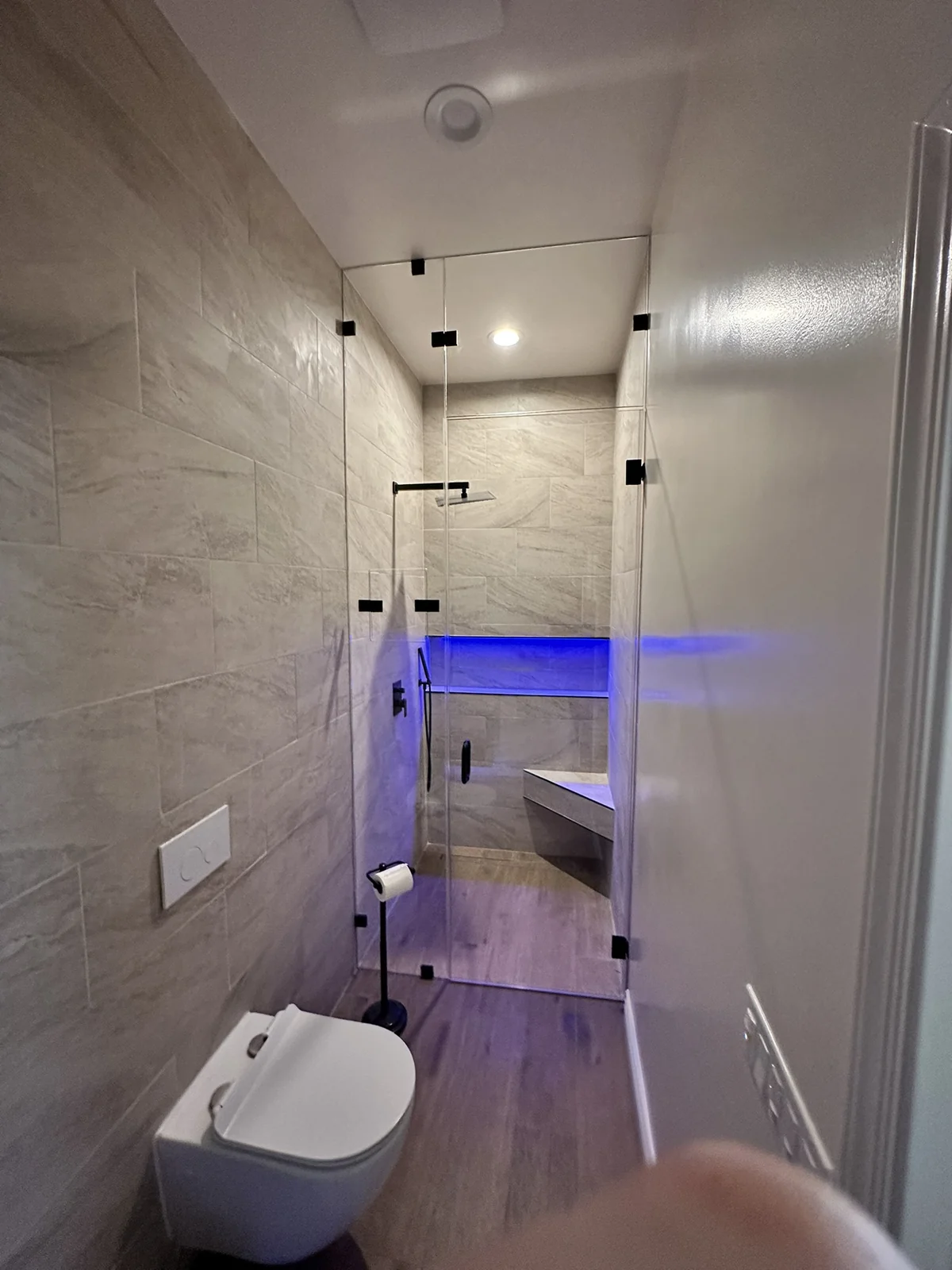 Narrow bathroom with a tiled shower, clear glass door, built-in bench and blue accent lighting