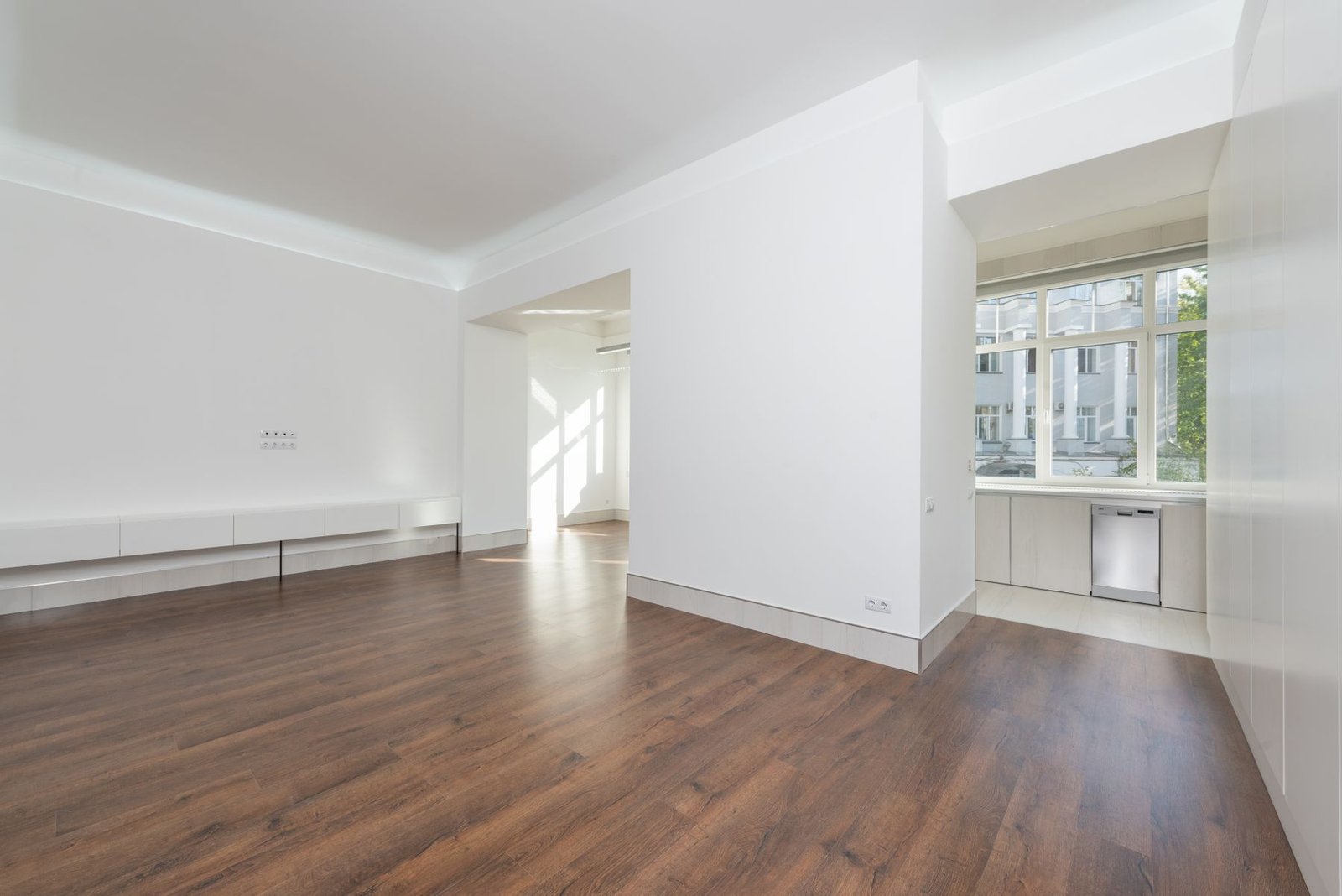 Empty living area with wood flooring, a wide doorway and an adjoining white kitchen