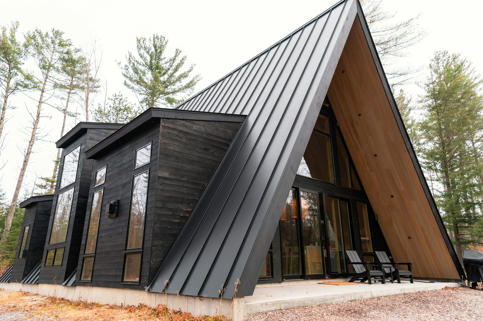 Timber-clad A-frame dwelling with a glazed entrance, chairs and surrounding trees