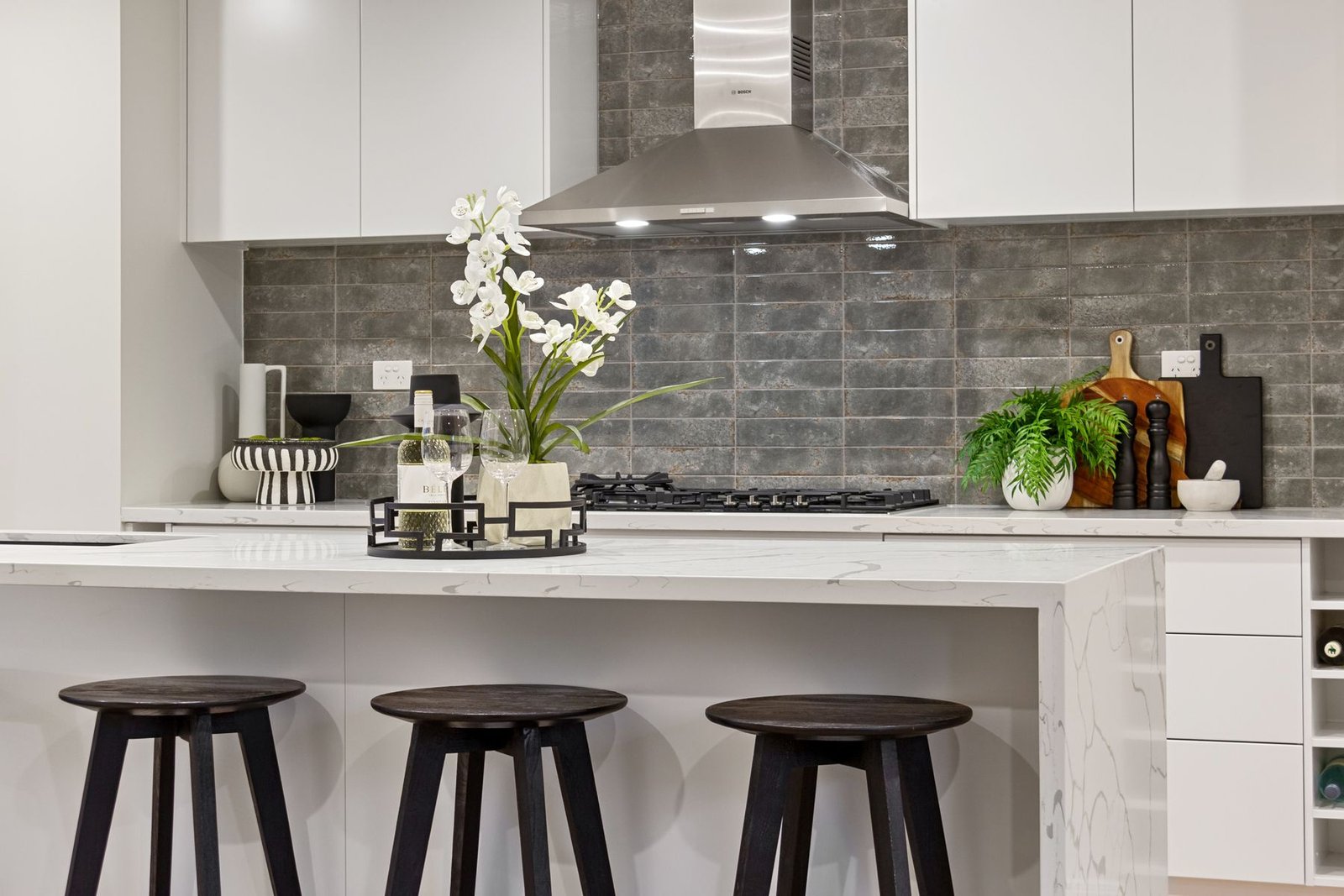 Kitchen island with three stools, white cabinets and a tiled cooking wall