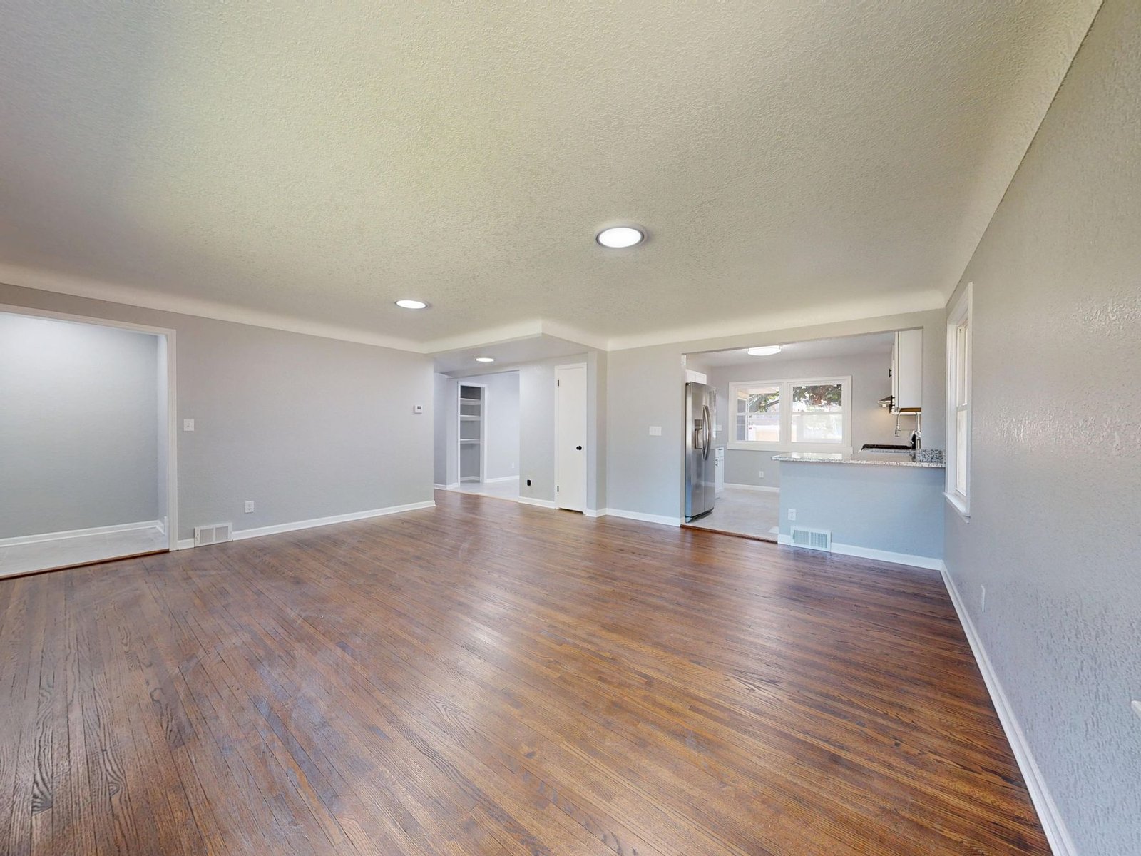Empty wood-floored living room connected to a kitchen and adjacent rooms.