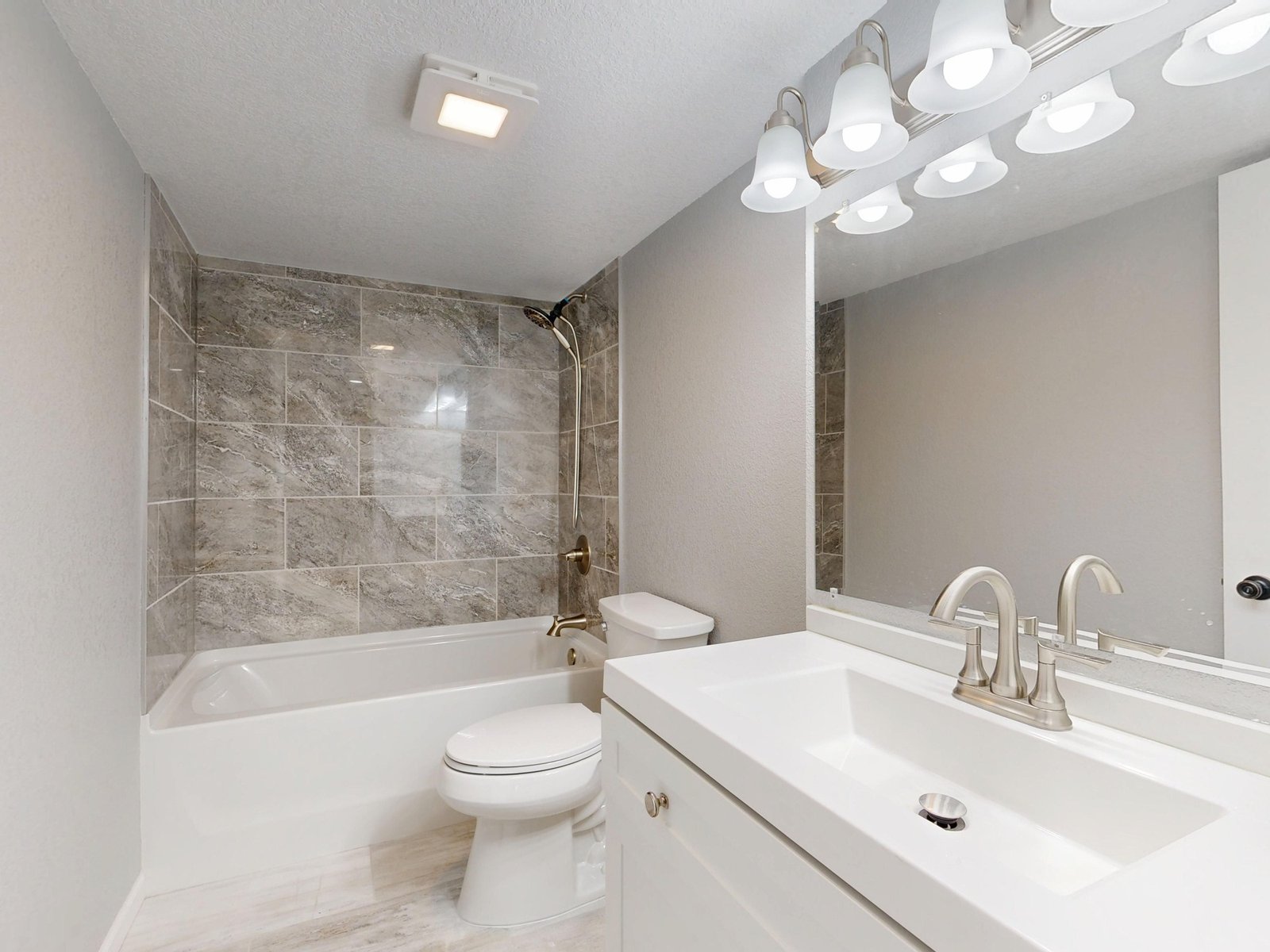 Bright bathroom with a ceiling fan and light unit above a tub-shower, beside a vanity and mirror