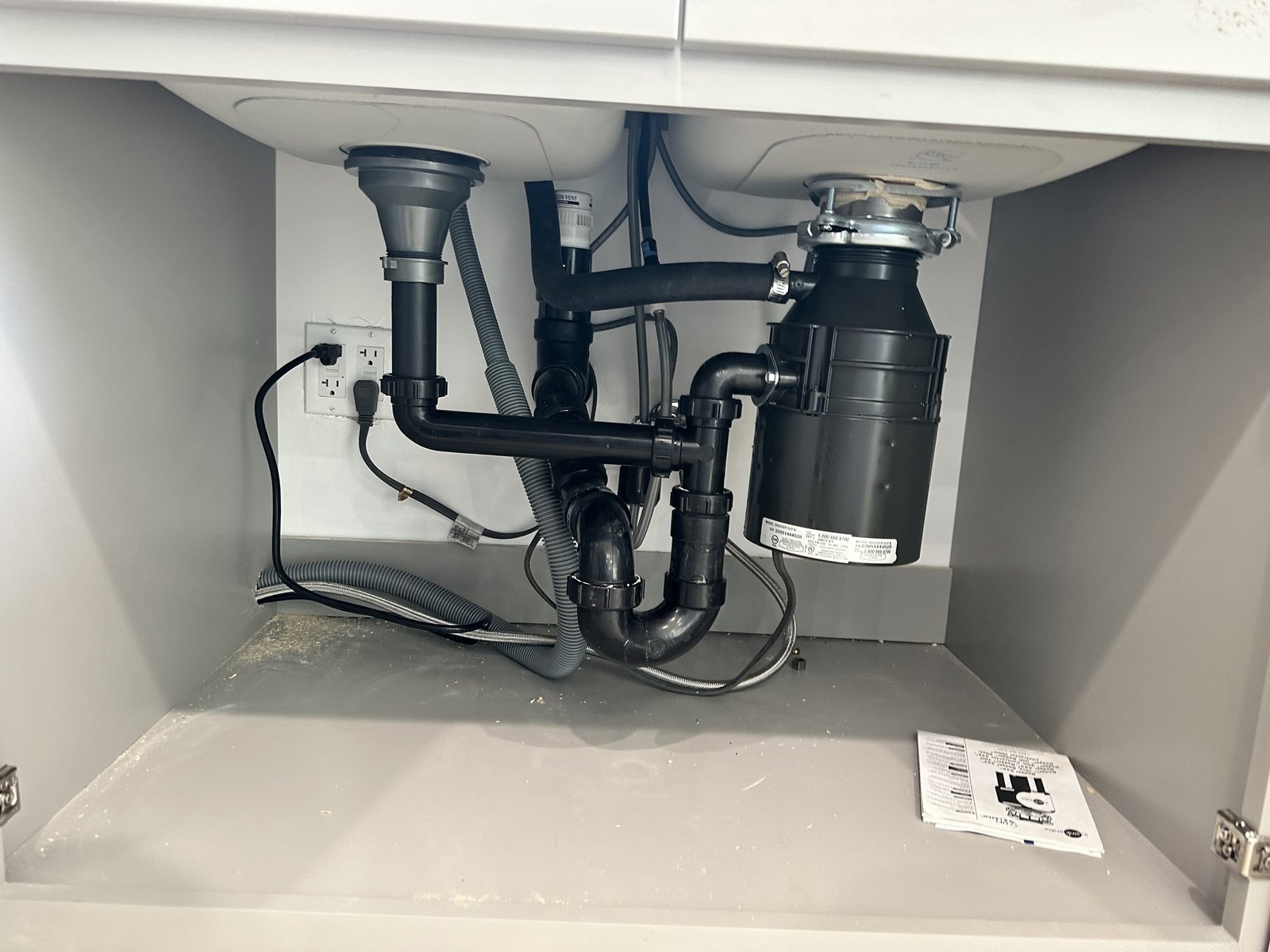 Open sink cabinet showing drain piping, disposal unit, hoses and electrical outlets