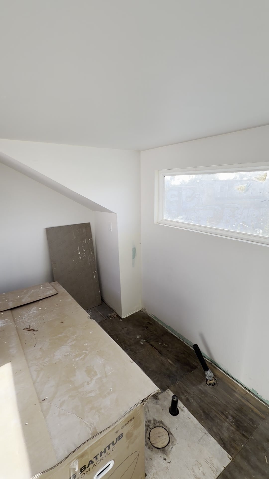 Unfinished white-walled room with a high window, exposed pipe stubs and a boxed bathtub
