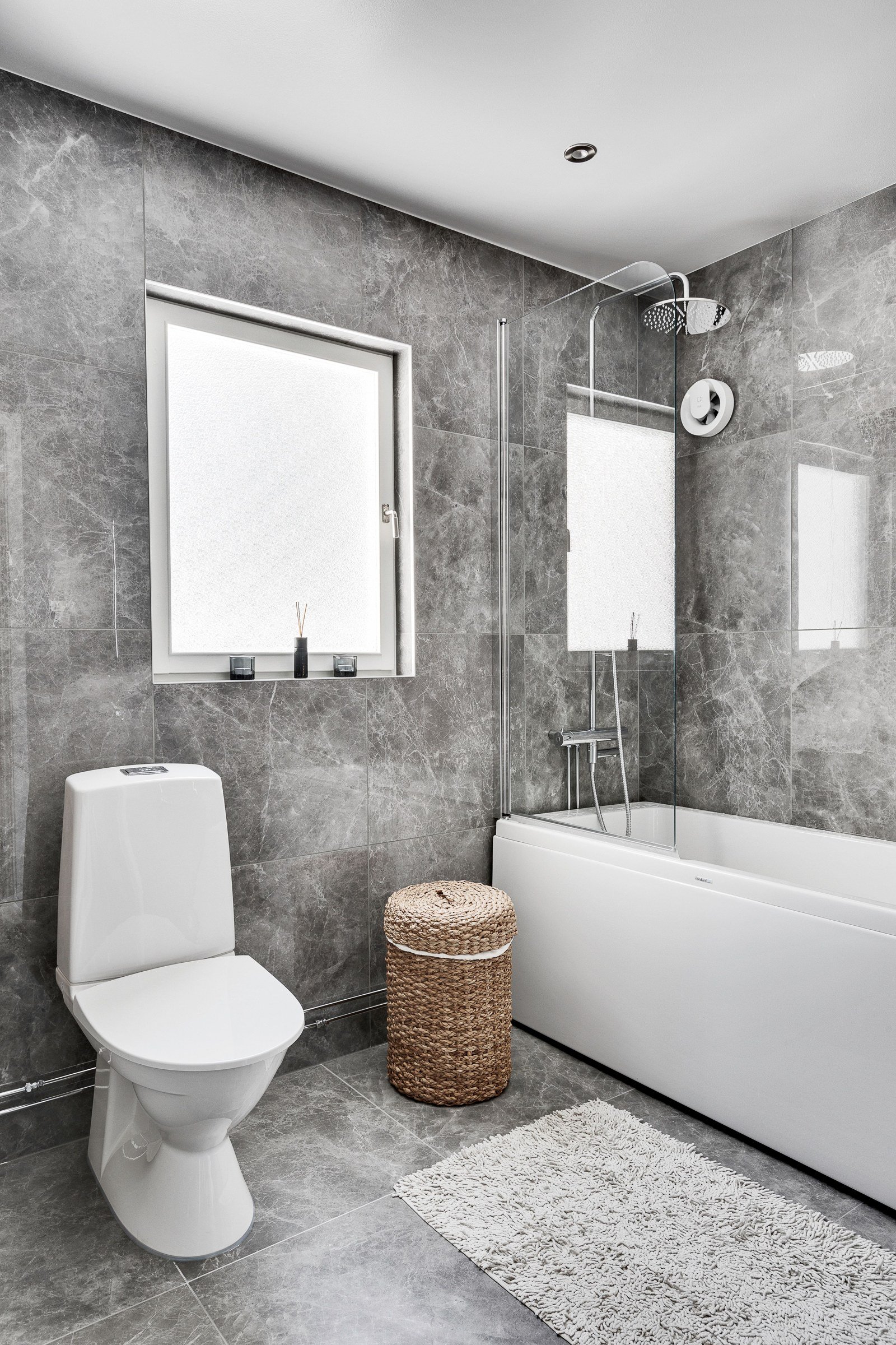 Gray veined bathroom tile surrounding a toilet, bathtub and glass shower screen