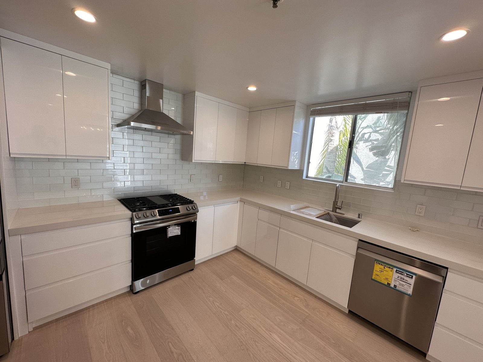 White kitchen with flat-front cabinets, stainless range, sink beneath a window and an L-shaped counter