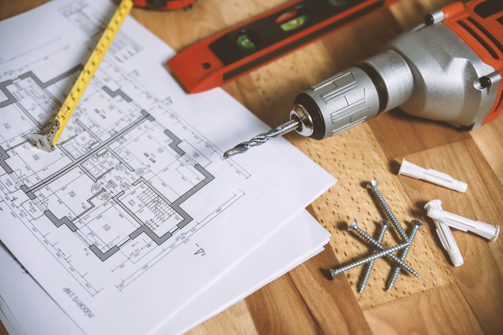 Printed residential floor plans beside a drill, tape measure, spirit level and fasteners on a wooden table