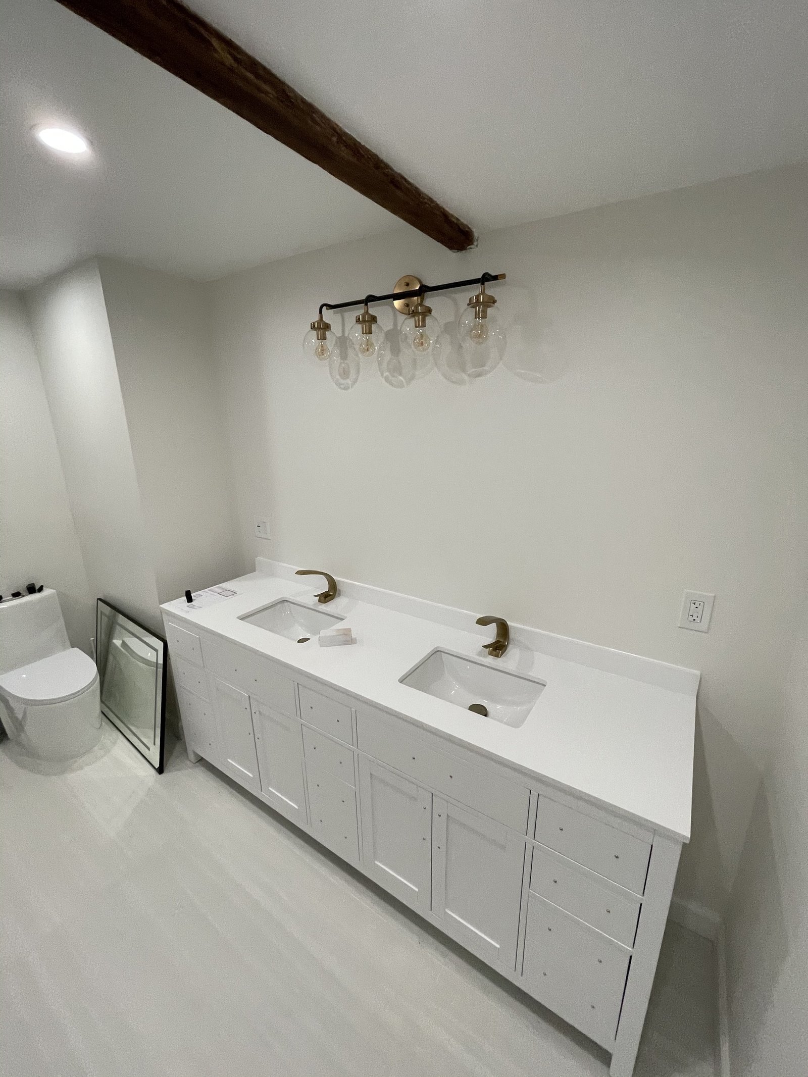 White double-sink vanity below a four-bulb wall light with mirrors resting against the wall
