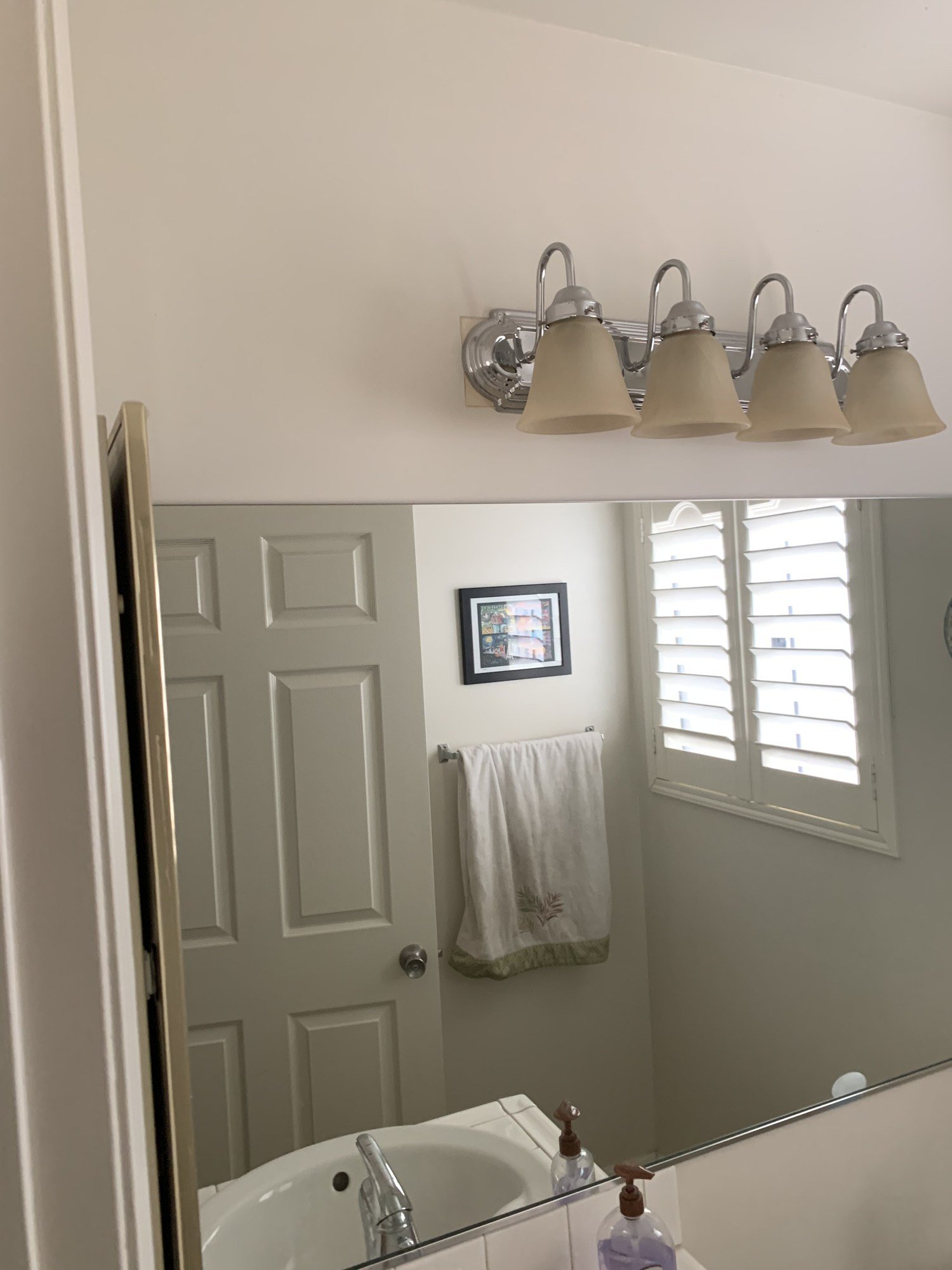 Bathroom mirror beneath a four-light fixture with a shuttered window reflected in the glass
