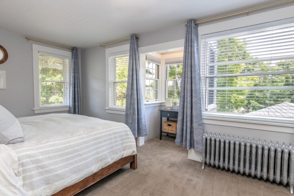Bedroom with a made bed, several windows, blue-gray curtains and a radiator