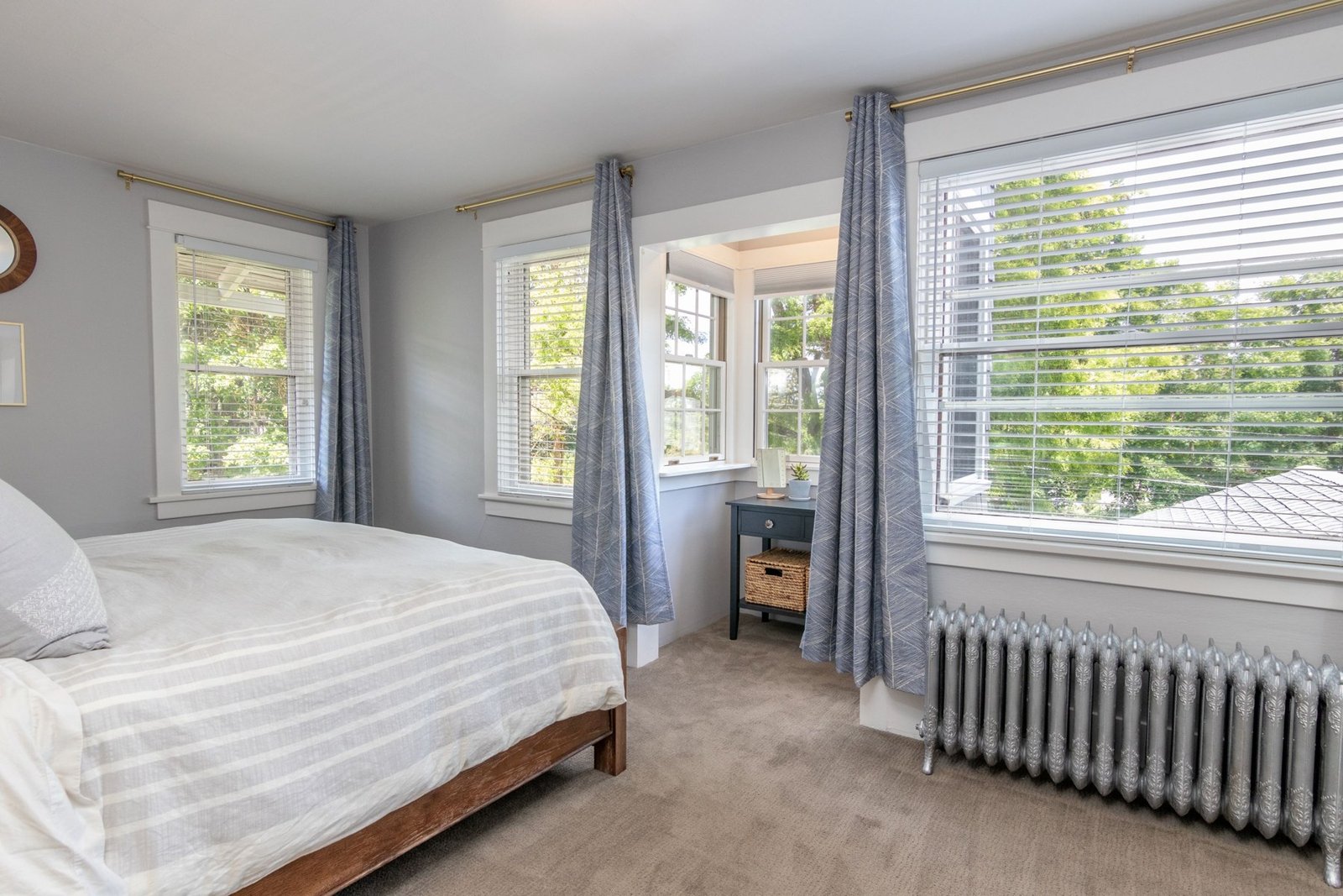 Bedroom with a made bed, several windows, blue-gray curtains and a radiator