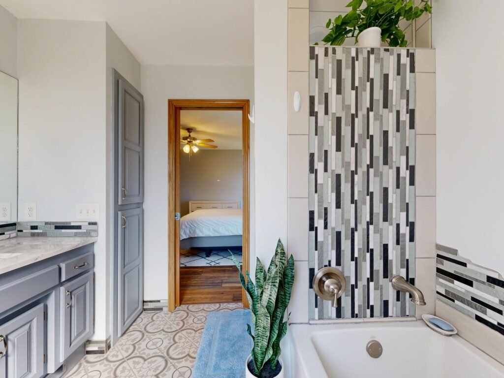 Bathroom with gray vanity and tiled tub surround looking through an open doorway toward a bedroom