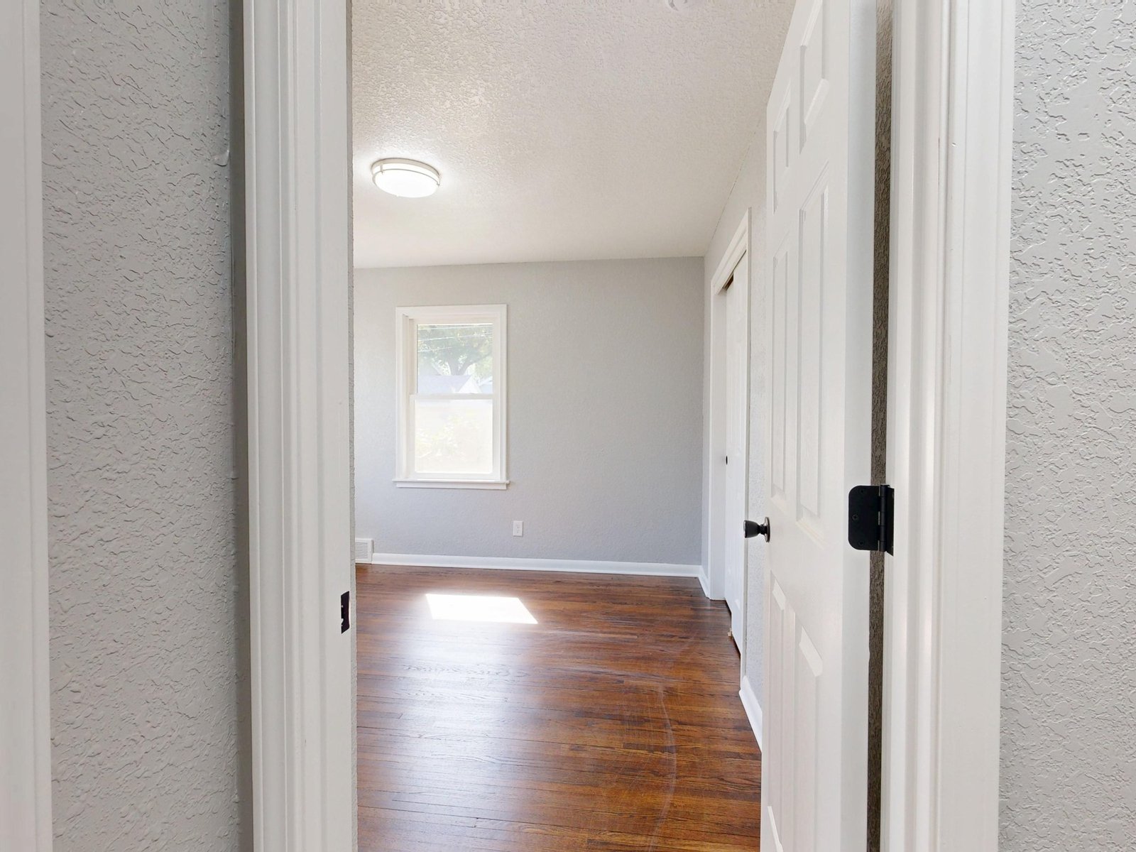 View through an open white door into an empty room with a wood floor, window and closet