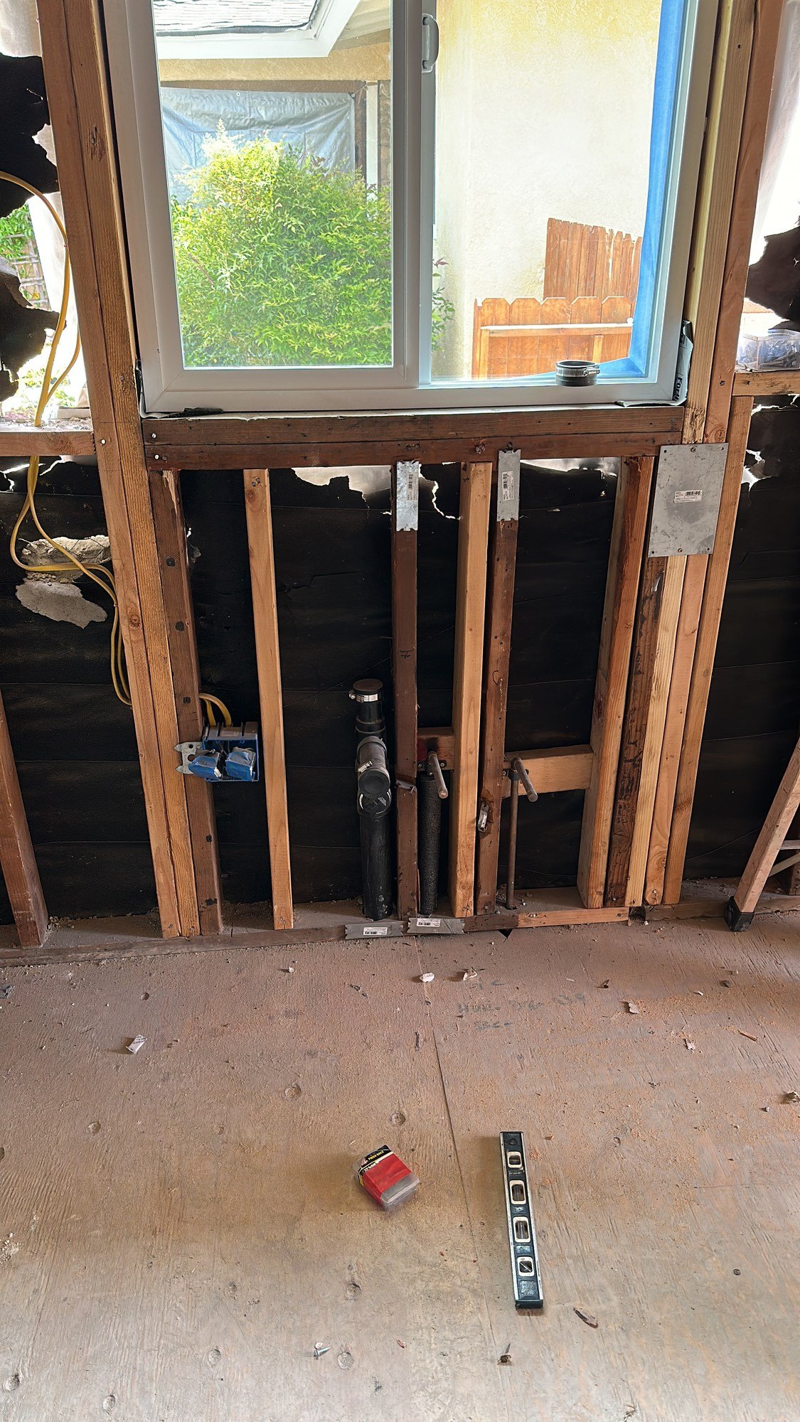 Exposed wood studs beneath a window with piping, an electrical box and a wood subfloor
